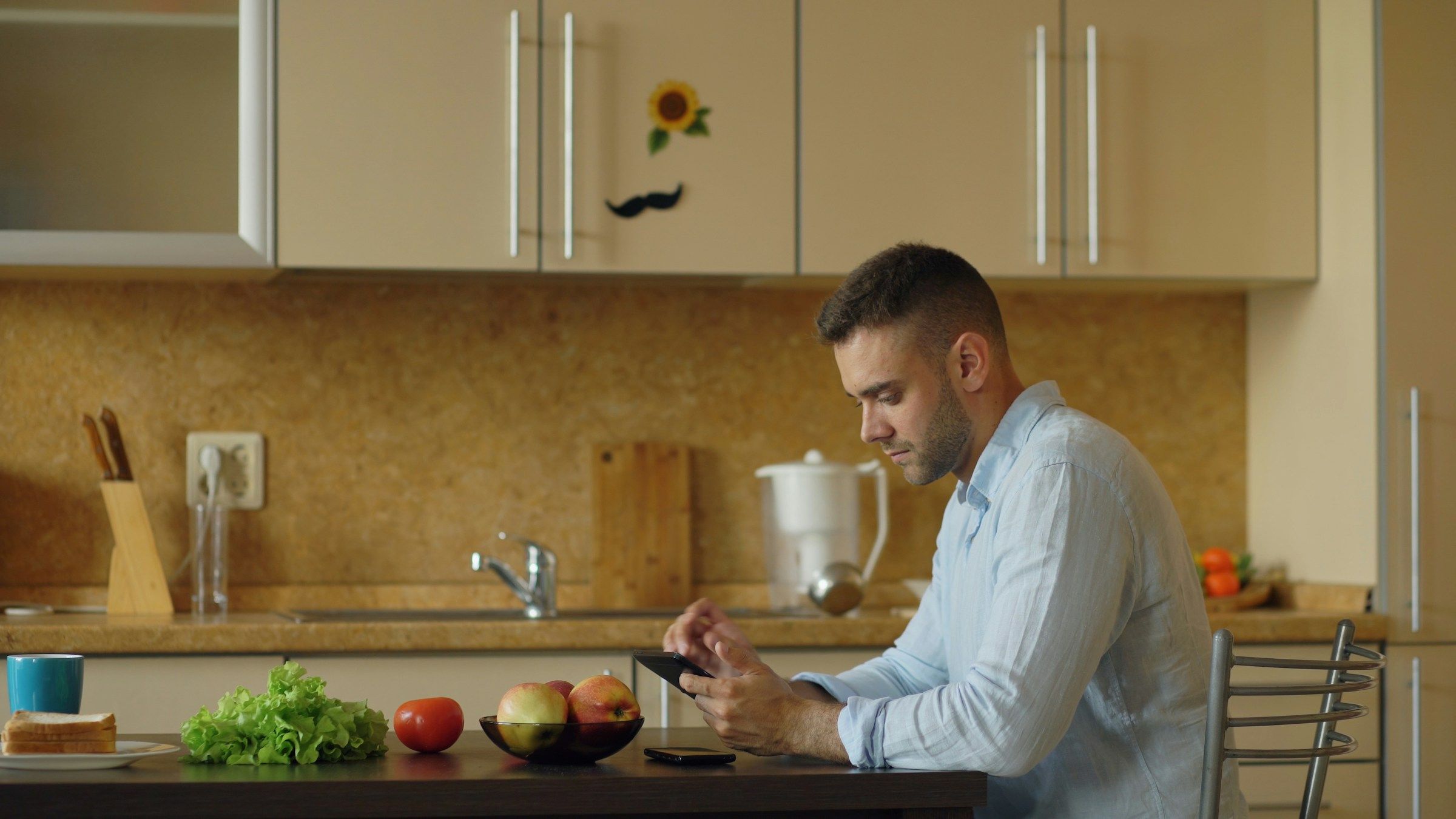 Un homme assis à la table de sa cuisine, en plein jour, consulte son téléphone.